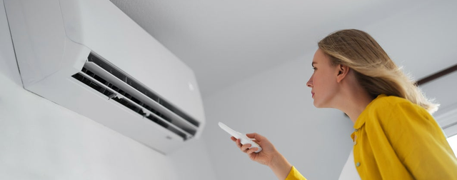 Kobieta w żółtej koszuli ustawia temperaturę pilotem, kierując go na biały klimatyzator ścienny w pokoju.