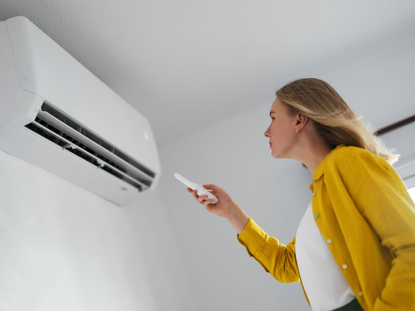 Kobieta w żółtej koszuli ustawia temperaturę pilotem, kierując go na biały klimatyzator ścienny w pokoju.