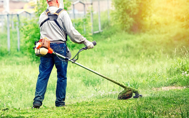 Mężczyzna w szelkach i rękawicach koszący trawnik kosą spalinową w słoneczny dzień.