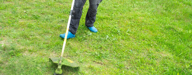 Osoba w spodniach roboczych kosząca trawę kosą spalinową w ogrodzie.