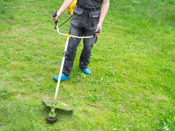 Osoba w spodniach roboczych kosząca trawę kosą spalinową w ogrodzie.