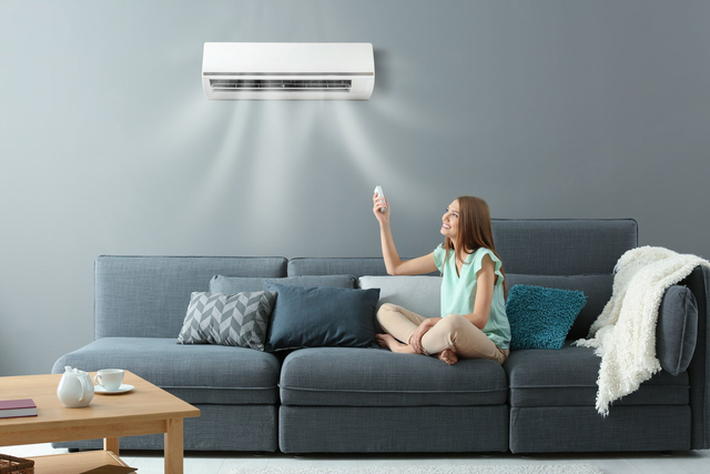 Uśmiechnięta kobieta siedzi na szarej kanapie w salonie i steruje klimatyzatorem ściennym za pomocą pilota.
