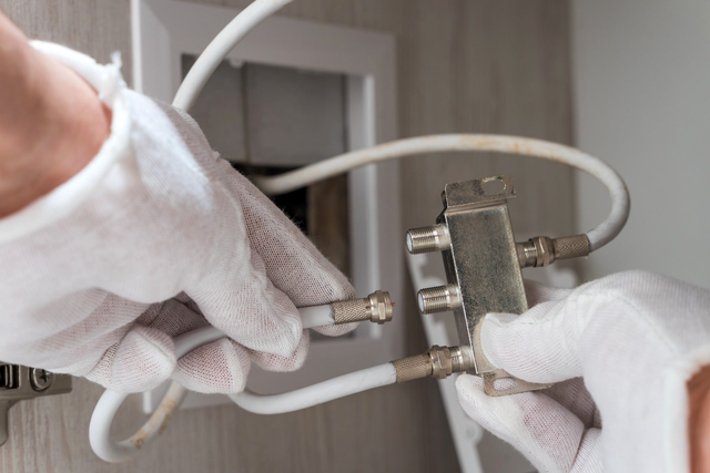 Dłonie w białych rękawiczkach przykręcają biały kabel antenowy z wtykiem F do metalowego rozgałęźnika sygnału.