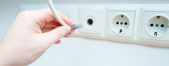 Dłoń podłączająca biały kabel antenowy z metalowym złączem do gniazda TV w białej ramce na ścianie.