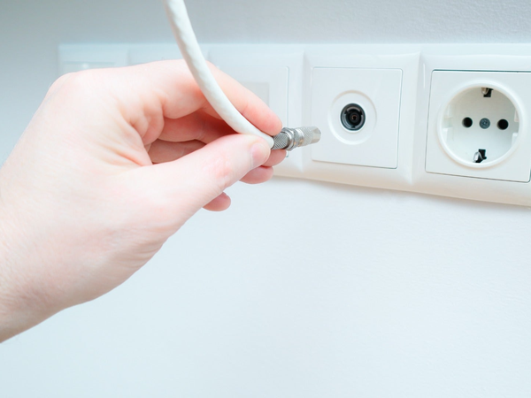 Dłoń podłączająca biały kabel antenowy z metalowym złączem do gniazda TV w białej ramce na ścianie.